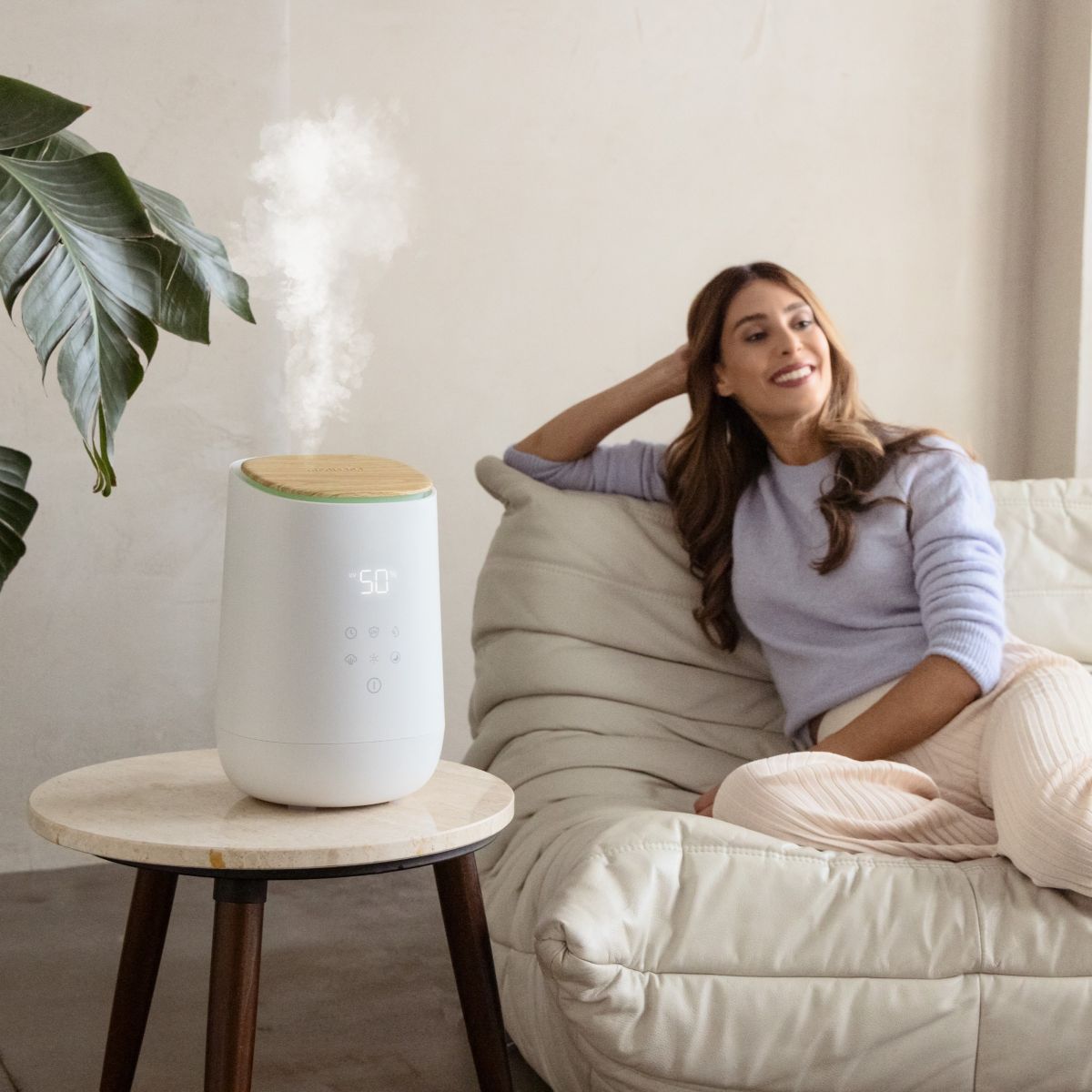 A modern humidifier releases visible mist on a side table in the living room while a woman relaxes on the sofa.